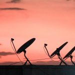 Fileira de antenas parabólicas de comunicação por satélite em um telhado, em silhueta contra o céu alaranjado do pôr do sol