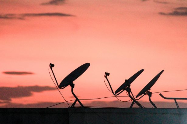 Fileira de antenas parabólicas de comunicação por satélite em um telhado, em silhueta contra o céu alaranjado do pôr do sol